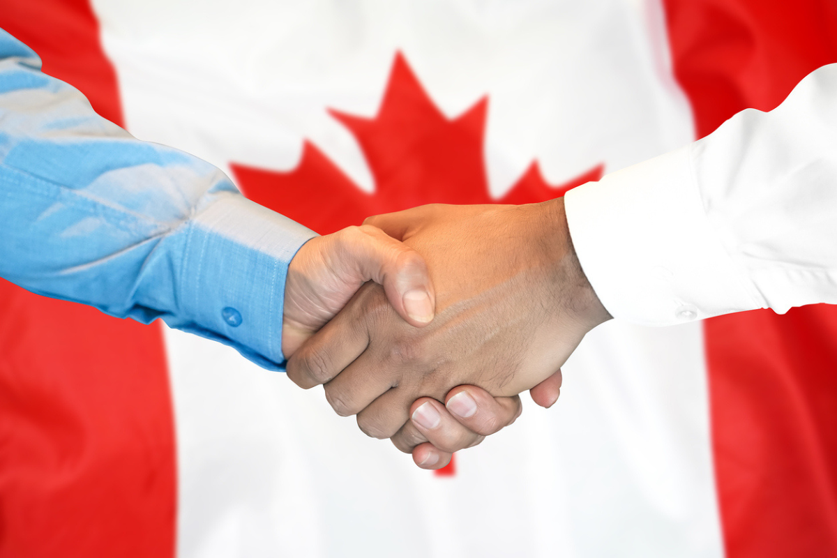 Close-up of two men in dress shirts shaking hands in front of the Canadian flag.