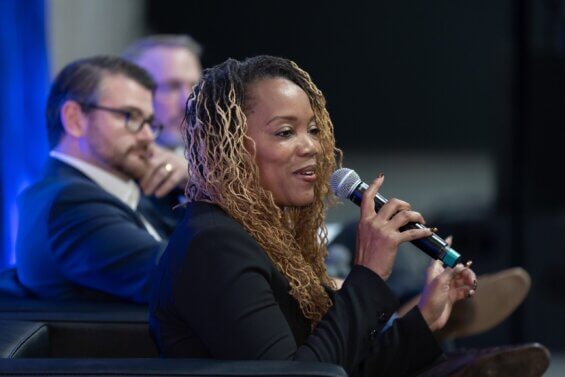 Close up of a Black woman looking out into a crowd and speaking into a microphone at a conference.