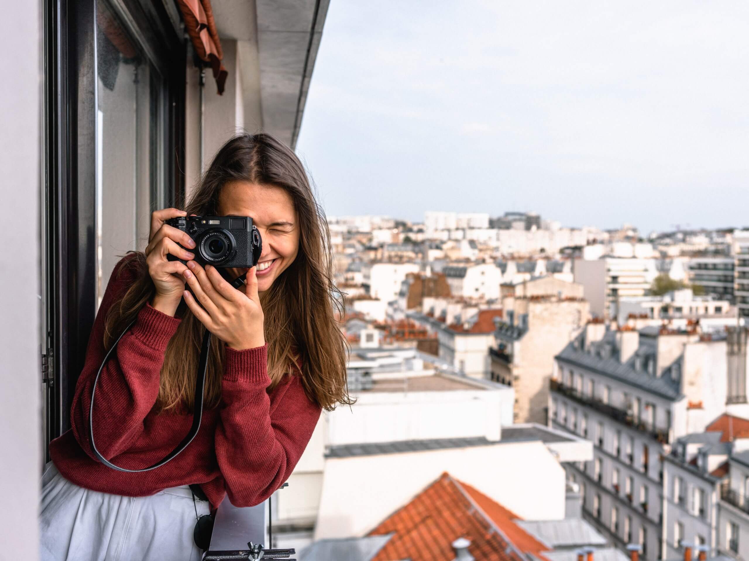 Female tourist smiling and taking a picture with her camera while leaning out of a window above Parisian rooftops.