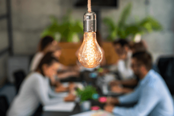 A bright light bulb shines in the foreground while a blurred group of of professionals sit at a conference table in the background
