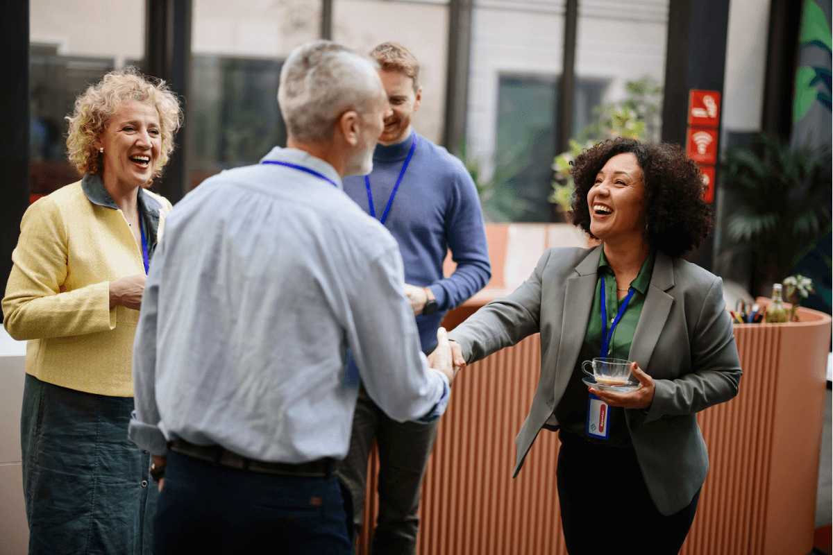 Older man shakes hands with smiling woman while standing in a group of people at a business conference.