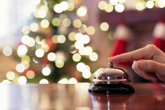Hand of guest ringing reception bell on desk of guesthouse, hotel at christmas time. Color shining garland on christmas tree on background. Travel concept.