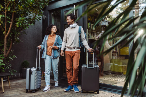 Black man and woman rolling suitcases outside of a modern glass apartment with lots of greenery.