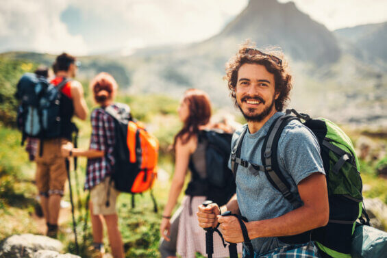 A small group of 20-somethings hike along a mountain trail, one man smiles at the camera.