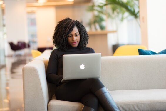 Black woman sits on a couch in a coworking space looking at her Macbook